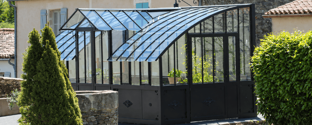 Serre Mirabeau LAMS noire sur embase de 87 cm, avec des plantes visibles à l’intérieur, dans un jardin au printemps.